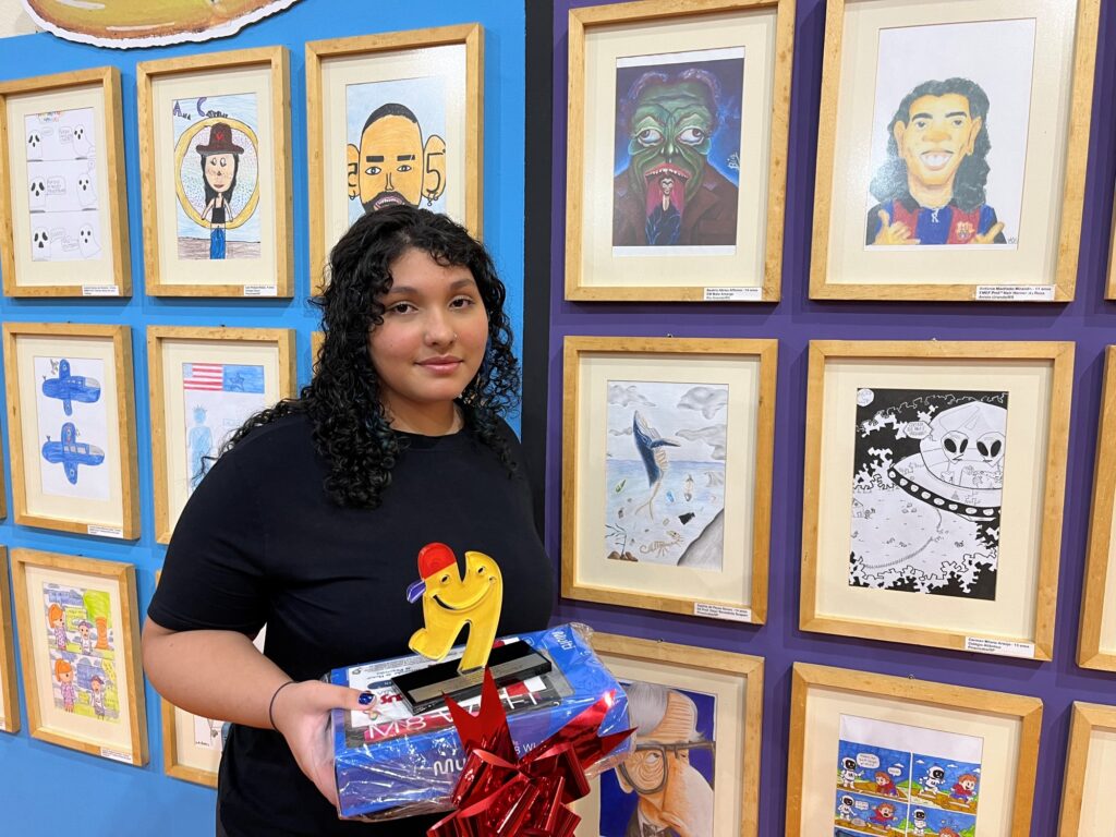 Sophia de Paula Nunes posa ao lado das obras expostas, segurando o seu troféu e prémio de 1.º lugar na categoria de 11 a 14 anos do Salãozinho de Humor.