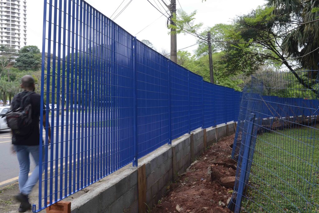 Um peão caminha junto a uma estrutura recém-instalada de gradil eletrofundido azul, com o antigo cercamento metálico ainda visível na área de terra ao lado.
