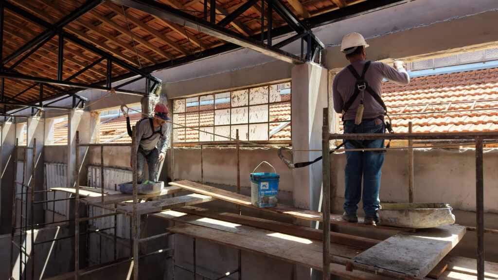 Dois trabalhadores da construção civil equipados com arneses de segurança trabalham num andaime interno, aplicando revestimento nas paredes superiores perto da estrutura do telhado.