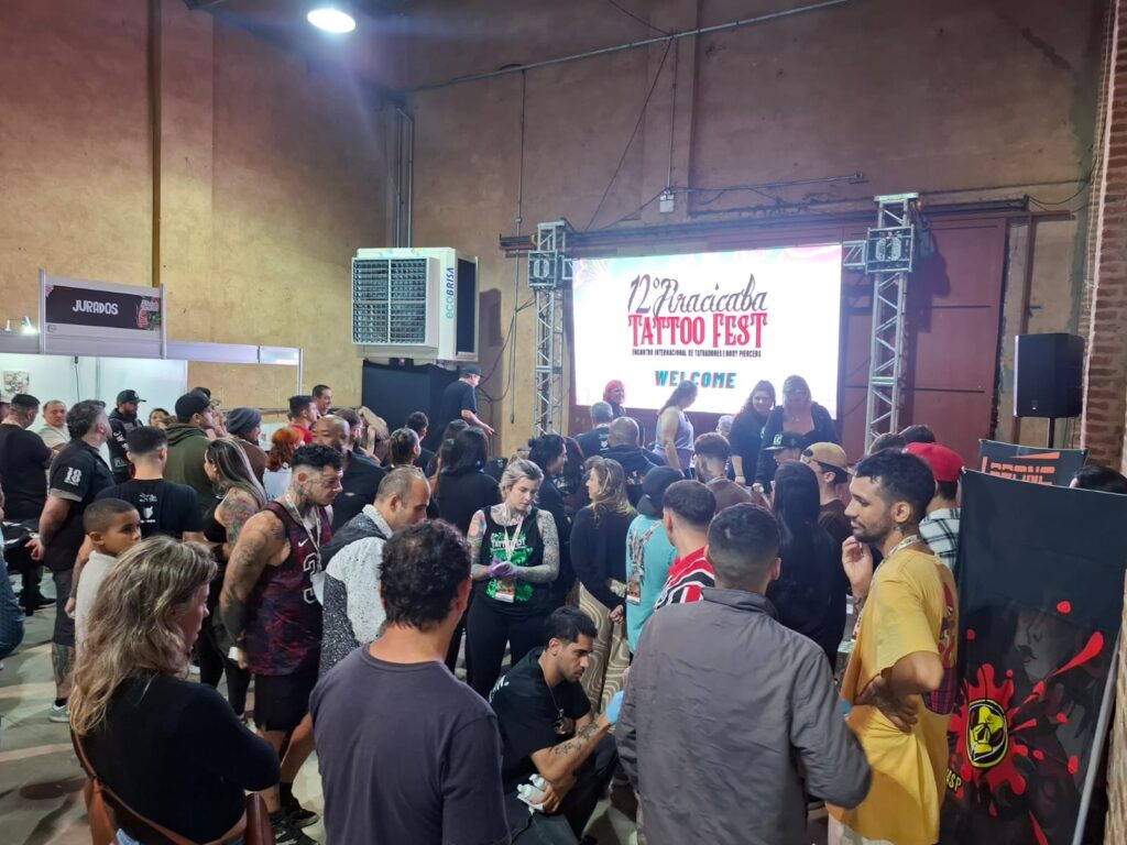 Público diversificado movimenta-se pelo galpão do Parque Engenho Central durante o evento Tattoo Fest, em frente ao painel do palco iluminado.