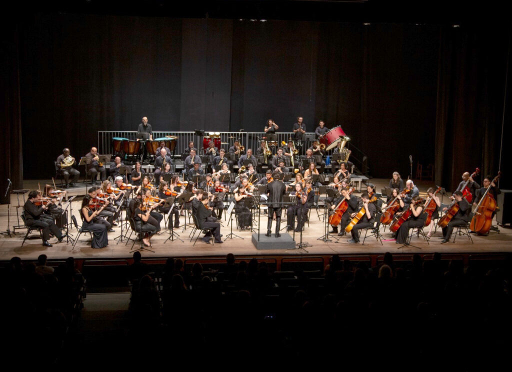 Vista geral do palco do Teatro Municipal Dr. Losso Netto durante uma apresentação da Orquestra Educacional de Piracicaba, sob a regência do maestro ao centro, rodeado pelos músicos com os seus respetivos instrumentos de cordas, sopro e percussão.