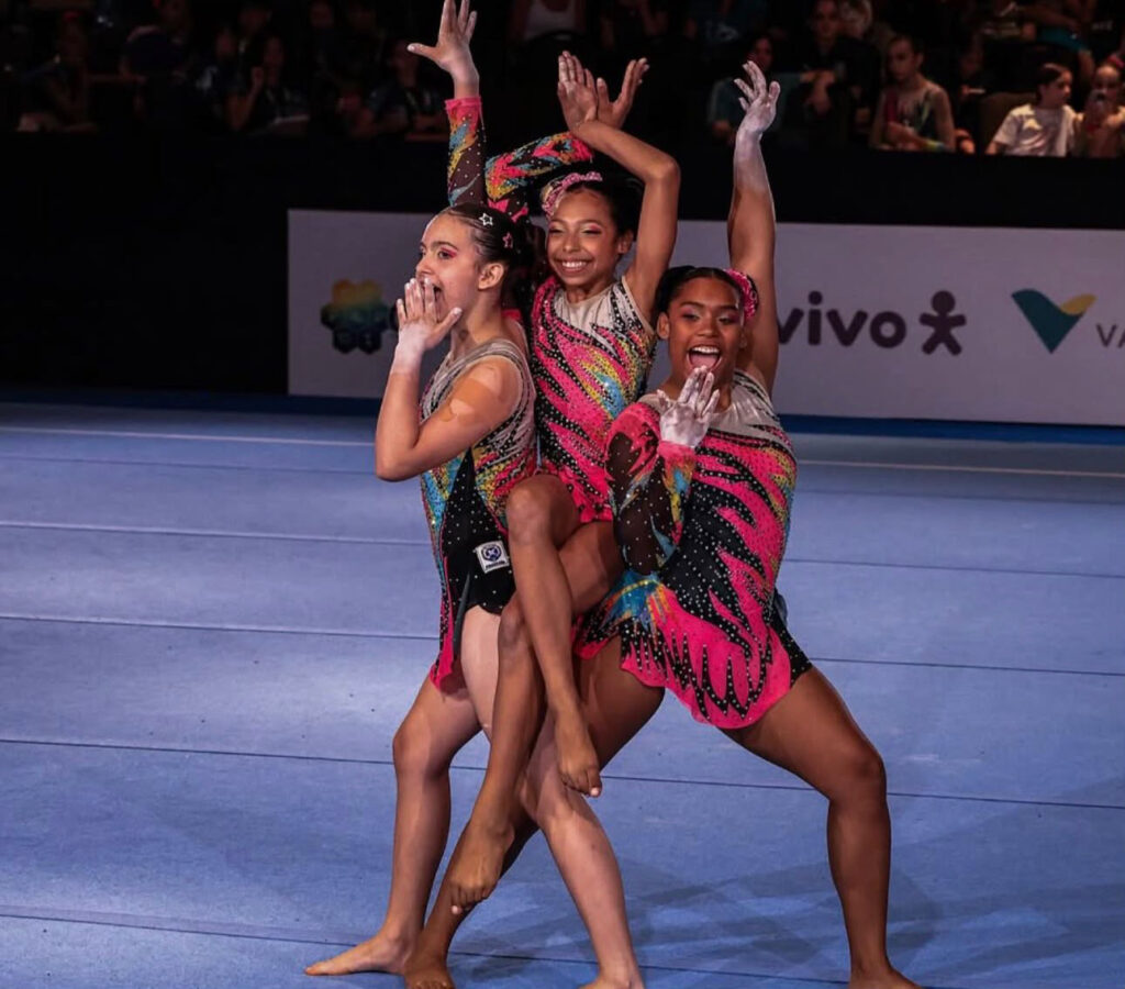 O trio formado por Sabrina de Andrade, Giovana Santin e Victória Olívio realiza uma pose acrobática expressiva durante a sua apresentação de ginástica.