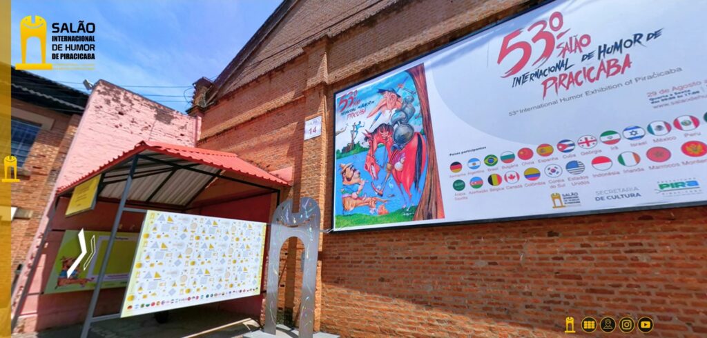 Entrada do Armazém 14, no Parque Engenho Central, em Piracicaba, espaço que recebe a exposição do 53º Salão Internacional de Humor de Piracicaba. A fachada é de tijolos aparentes, com uma grande faixa de divulgação do evento na parte superior direita. O painel apresenta o número 53º, o nome do Salão Internacional de Humor de Piracicaba e uma ilustração colorida com personagens e elementos de humor gráfico. Também é possível observar as bandeiras dos países participantes. À esquerda, encontra-se a entrada da exposição, protegida por uma cobertura de telhas vermelhas.