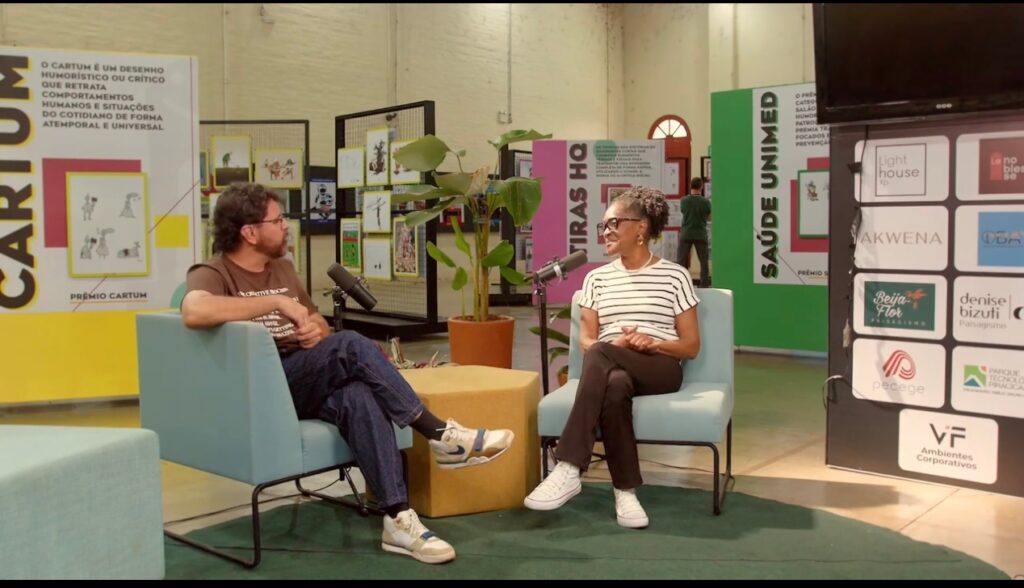Zetti Araújo, primeira diretora do Salão Internacional de Humor de Piracicaba, sentada numa poltrona em frente a um apresentador durante a gravação do videocast No Meio do Salão, num espaço de exposição decorado com painéis informativos e obras de cartoon.