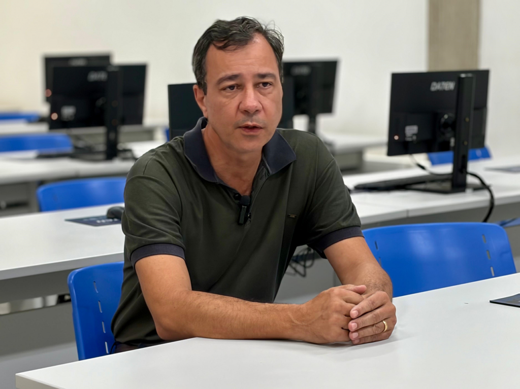 O professor Clerivaldo Roccia está sentado numa mesa, com as mãos entrelaçadas, concedendo uma entrevista dentro do laboratório de informática da EEP, rodeado por monitores e bancadas de estudo.